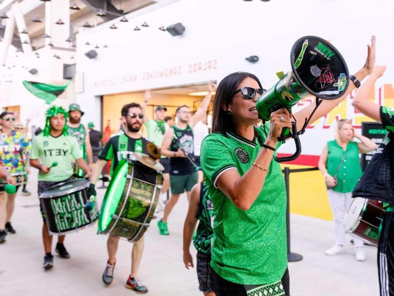 图为一群热情的粉丝, wearing green and black attire with "YETI" logos, celebrating and marching with drums and a megaphone inside a stadium. 场面很热闹。, with one person leading chants through the megaphone while others play drums and high-five. The overall atmosphere is energetic and festive, indicating a sports event or rally.